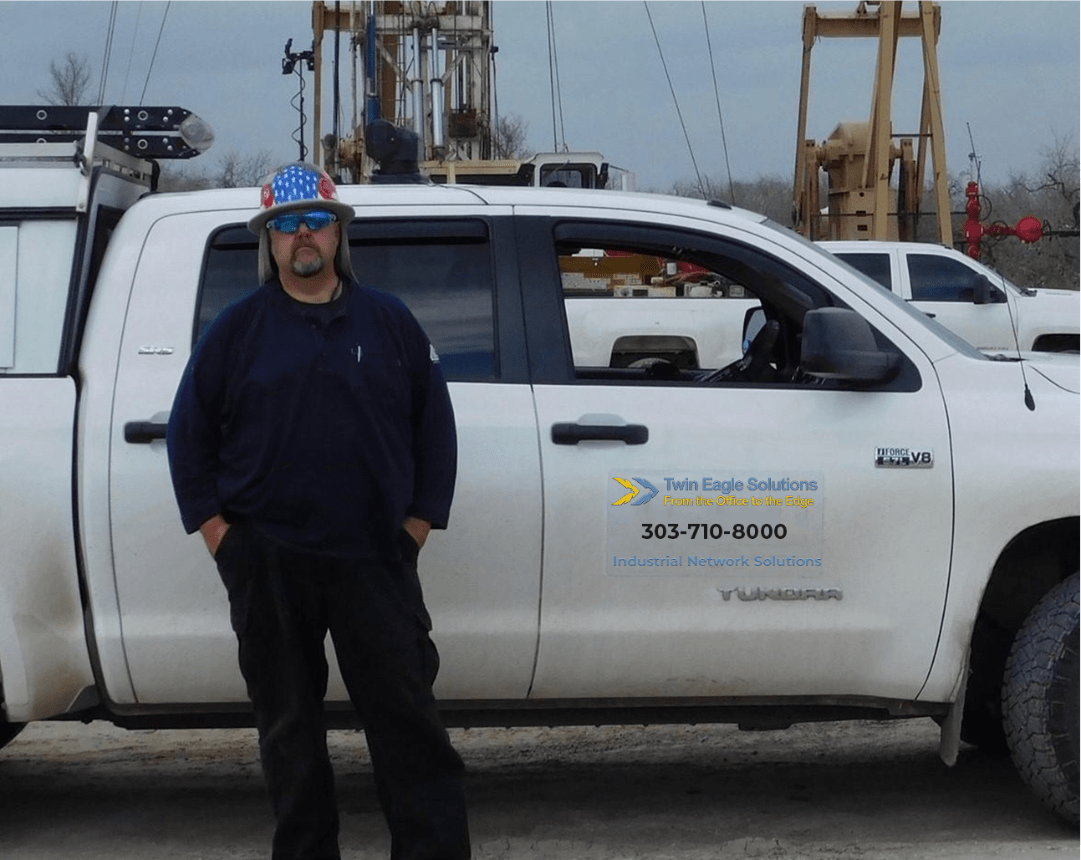 Twin Eagle field engineer standing beside a Twin Eagle Solutions service truck on an active oil and gas pad with a drilling rig and pumpjacks behind