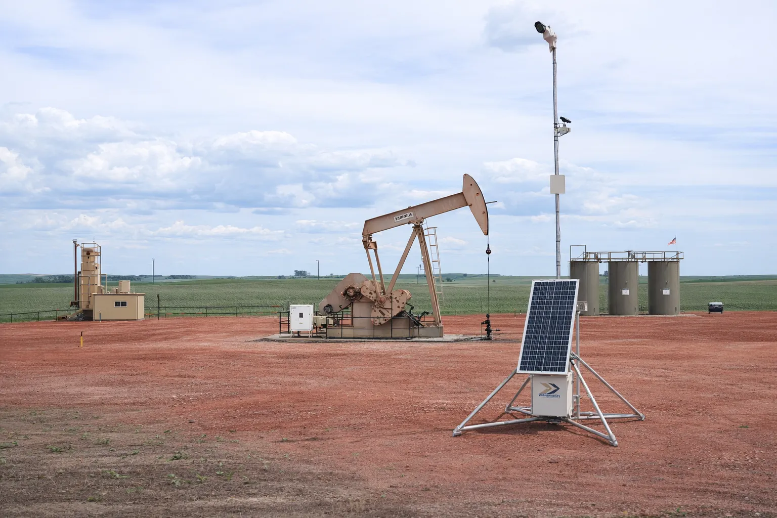 Solar-powered Remote Pumper skid with pole-mounted camera deployed beside a pumpjack and tank battery on a North Dakota well pad