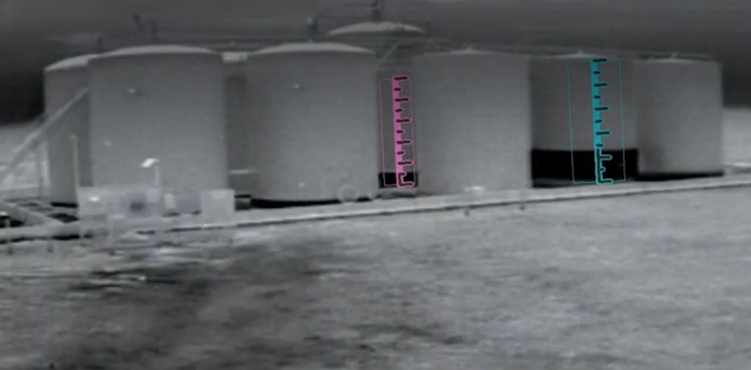 Thermal camera frame of a multi-tank battery with VisionAery digital rulers overlaid in pink and cyan — calibration ticks pegged to tank reference features, with the liquid line readable against the ruler