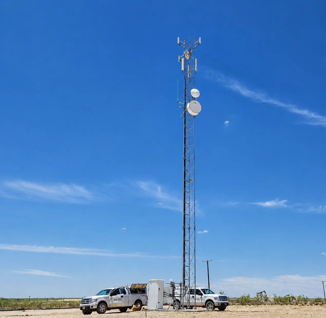 Twin Eagle Solutions service trucks at the base of a communications tower carrying cellular, microwave dish, and narrowband antennas at a remote oilfield site